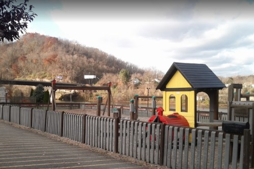 A playground with swings and a playhouse, set against a backdrop of a wooded hill under a cloudy sky.
