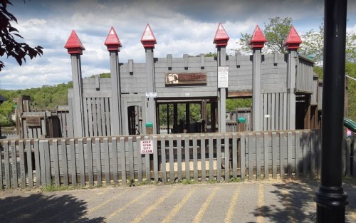 A wooden play structure with red-tipped towers, surrounded by a fence and trees under a cloudy sky.