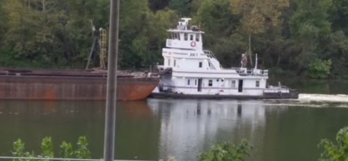 A tugboat navigates a river, pushing a large barge alongside lush green trees on the shore.