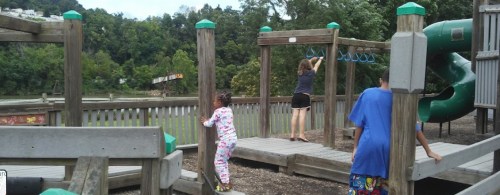 Children playing on a wooden playground with a slide and climbing structure, surrounded by greenery.