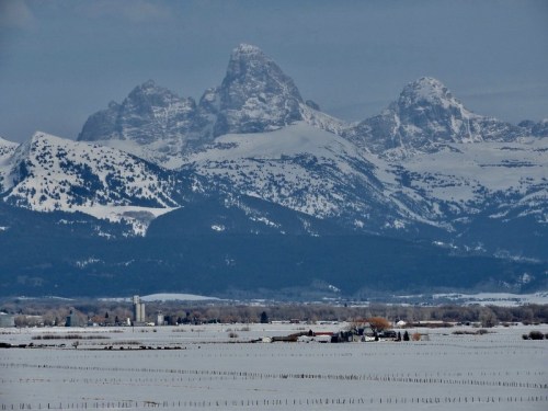 Snow-covered mountains rise above a flat, white landscape with a small farm in the foreground.