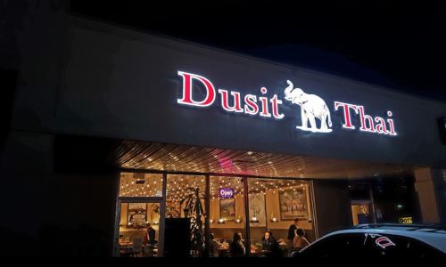 Exterior of Dusit Thai restaurant at night, featuring a lit sign and warm interior ambiance with diners inside.