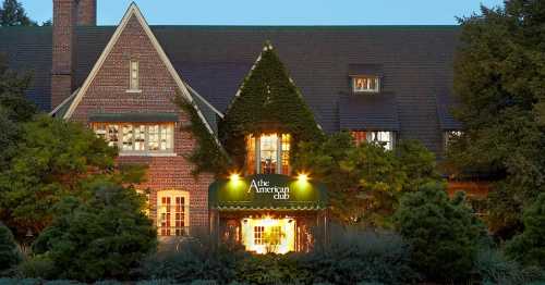 A charming brick building with ivy, featuring a green awning that reads "The American Club," surrounded by trees at dusk.