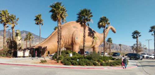 A large dinosaur statue surrounded by palm trees, with people walking nearby and mountains in the background.