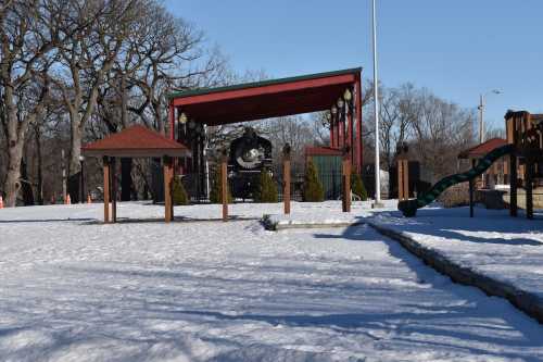 A snowy park scene featuring a vintage steam locomotive under a covered structure, with playground equipment nearby.