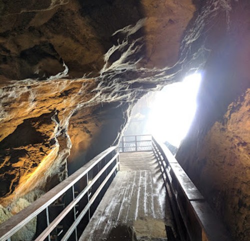 A wooden walkway leads through a rocky cave, illuminated by light at the end, creating a mysterious atmosphere.