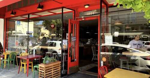 A vibrant café with red exterior, open doors, and colorful outdoor seating. People are inside and outside enjoying the space.