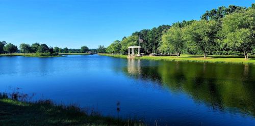 A serene lake surrounded by lush greenery and a gazebo, reflecting the clear blue sky.