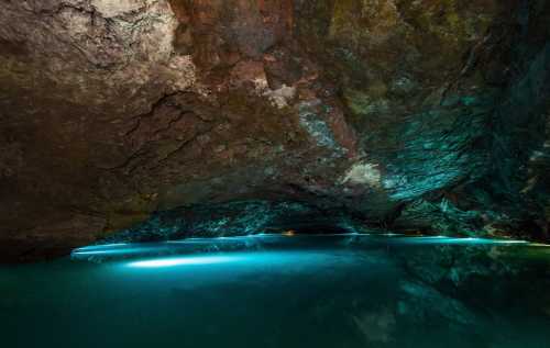 A serene underground cave with a shimmering blue water surface and textured rock formations above.