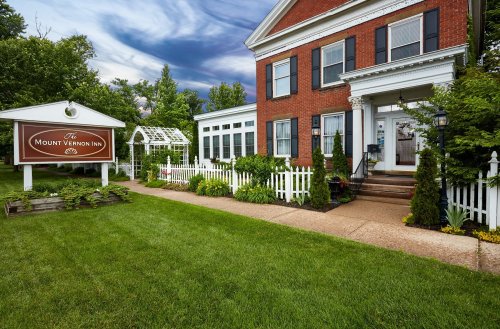 A charming brick inn with white trim, surrounded by lush greenery and a well-kept lawn, under a cloudy sky.