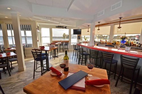 A cozy restaurant interior with tables, a bar, and large windows overlooking a waterfront view.