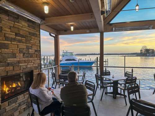 A couple sits on a patio by the water, enjoying a sunset view with a boat in the distance and a cozy fireplace nearby.