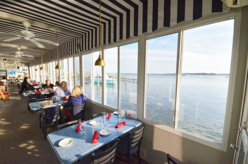A waterfront restaurant with striped ceilings, tables set for dining, and a view of the water and boats outside.