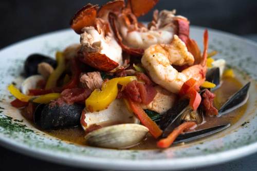 A colorful seafood dish featuring lobster, shrimp, mussels, and vegetables in a savory broth on a white plate.