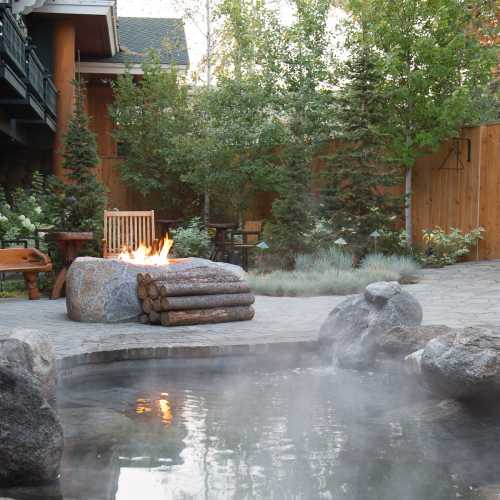 A cozy outdoor space featuring a stone fire pit, wooden chairs, and lush greenery surrounding a steaming hot tub.