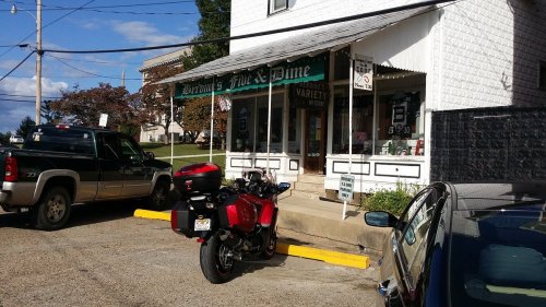 A small store with a porch, surrounded by parked cars and a motorcycle, under a clear blue sky.
