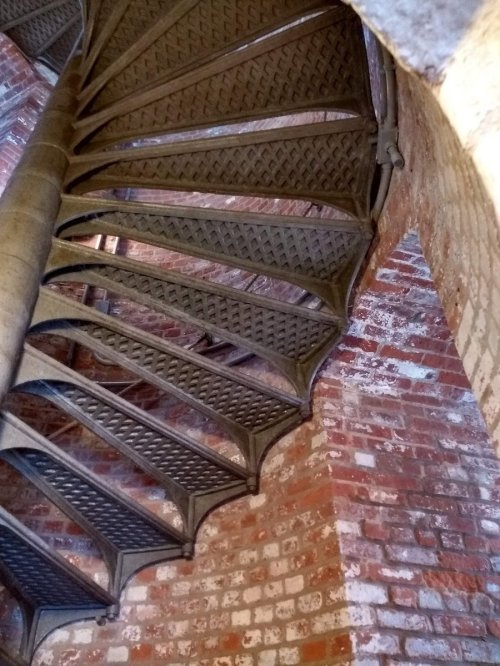 A spiral staircase with metal steps, viewed from below, against a backdrop of red brick walls.
