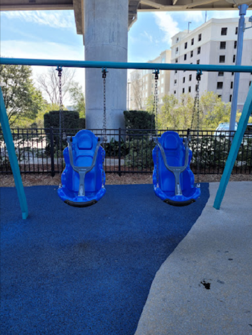 Two blue swings hanging from a frame, set against a backdrop of trees and buildings under a clear sky.