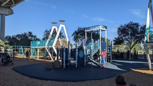 A playground with blue equipment, under a clear sky, featuring slides and climbing structures, with people nearby.