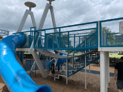 A blue slide and climbing structure at a playground, with children and adults playing in a sunny outdoor setting.