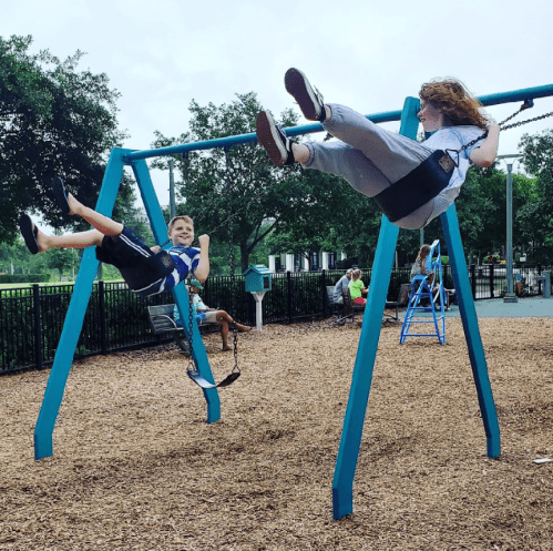 Two children swing on a playground, smiling and enjoying their time on swings surrounded by trees and other kids.