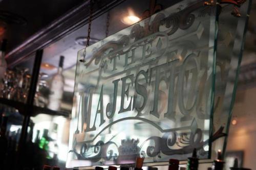 A decorative glass sign reading "The Majestic" hangs in a bar, with blurred bottles and glasses in the background.