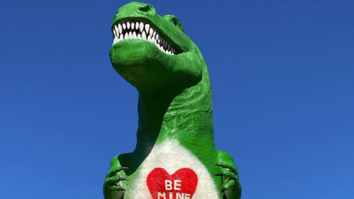A large green dinosaur statue with a heart that says "BE MINE" against a clear blue sky.