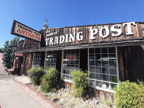A rustic trading post building with a sign reading "Trading Post Antiques" and large windows under a clear blue sky.