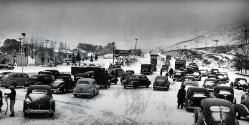 A snowy scene with vintage cars and trucks on a road, near a motel sign, surrounded by hills and trees.