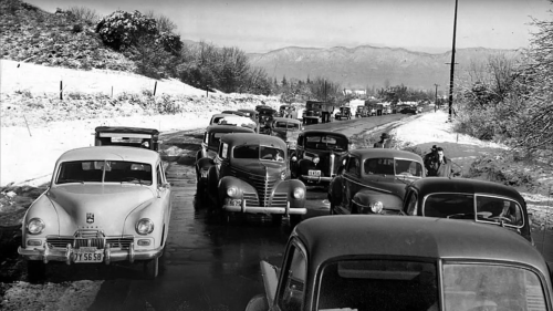 A vintage black-and-white photo of a snowy road with a line of classic cars and mountains in the background.
