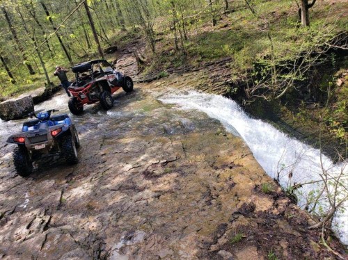 Two ATVs parked near a waterfall in a lush, green forest setting. Water cascades over rocky terrain.
