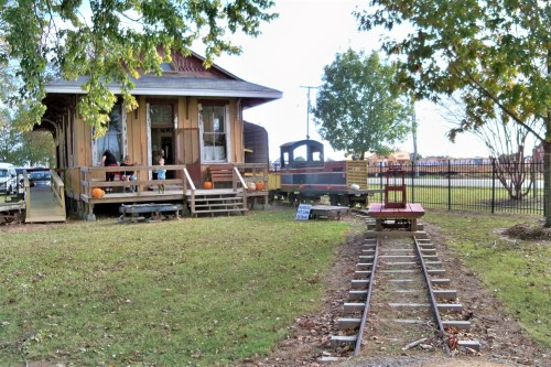 A wooden train station with a small train and tracks, surrounded by trees and grassy areas.