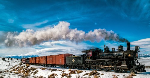 A vintage steam locomotive pulls a train of red freight cars through a snowy landscape under a blue sky.