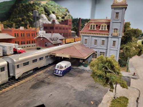 A miniature train station with a vintage train and a small blue and white vehicle parked outside. Scenic backdrop included.