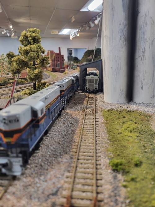 A model train scene featuring two locomotives on tracks, passing through a tunnel with trees and buildings in the background.