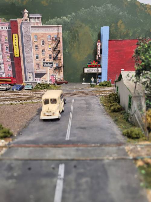A miniature scene featuring a road with a vintage car, buildings, and a train track in a model town setting.