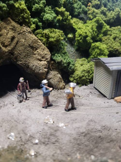 Three miniature figures walking near a small building, surrounded by lush greenery and rocky terrain.