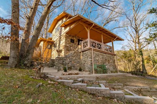 A charming stone cottage with a wooden porch, surrounded by trees and a grassy area, under a clear blue sky.
