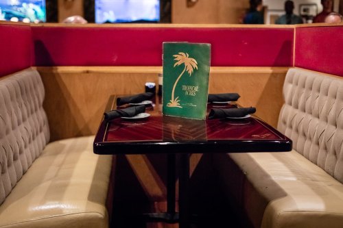 A cozy restaurant booth with a green menu titled "Tropical Isles" on a polished table, surrounded by red and beige decor.