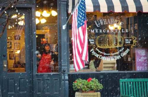 A cozy mercantile shop with an American flag, snow falling outside, and a person in an apron visible through the window.