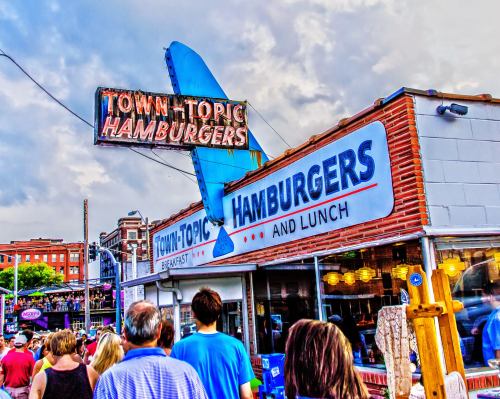 A bustling hamburger restaurant with a retro sign, people walking by, and a cloudy sky in the background.