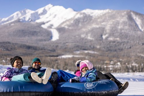 Four children sit on snow tubes, smiling against a backdrop of snowy mountains and a clear blue sky.