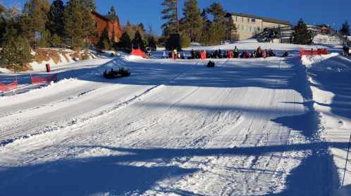 A snowy hill with people sledding down, surrounded by trees and buildings under a clear blue sky.