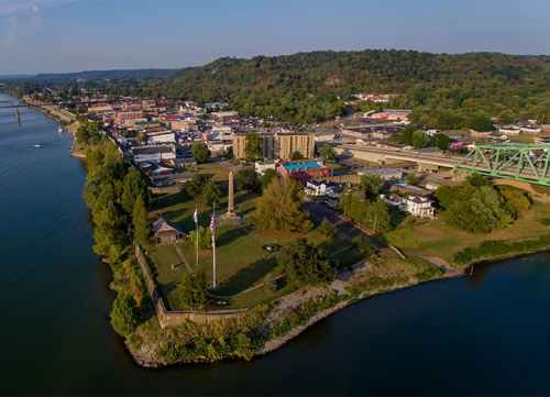 Aerial view of a riverside town with buildings, greenery, and a bridge in the background under a clear blue sky.