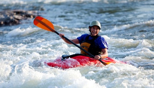 A person in a red kayak paddles through whitewater rapids, smiling and wearing a helmet and life jacket.