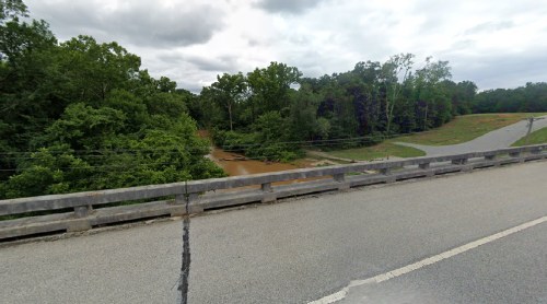 A view from a bridge overlooking a muddy river surrounded by lush green trees and a winding road.