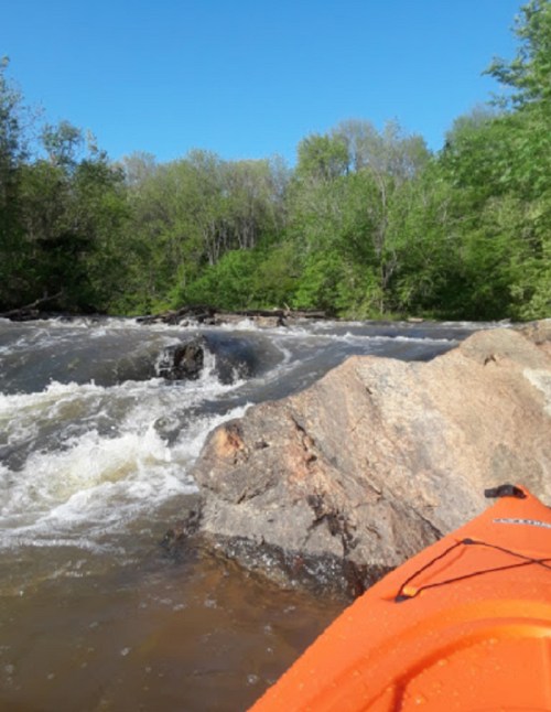 A vibrant orange kayak near rocky river rapids, surrounded by lush green trees under a clear blue sky.