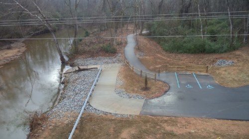A winding path alongside a river, with a parking area featuring accessible spaces and bare trees in the background.