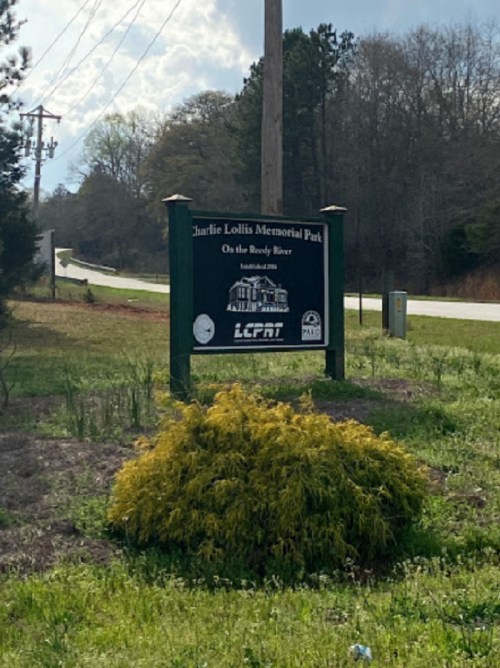Sign for Charlie Lollis Memorial Park, located on the Rocky River, with surrounding greenery and a road in the background.