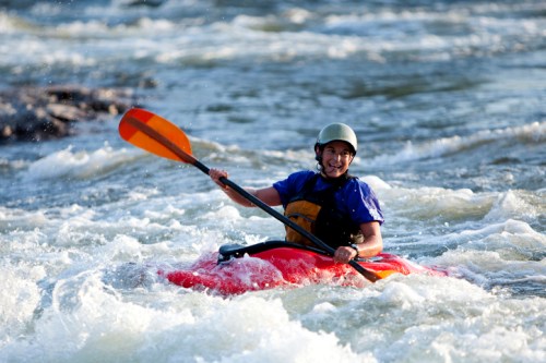 A person in a kayak navigates through whitewater rapids, smiling and paddling with an orange paddle.